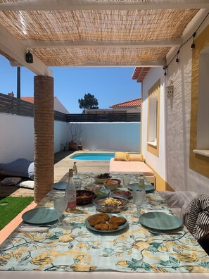 An outdoor dining area with a pool in the background, a table set with various dishes, and a pergola overhead.
