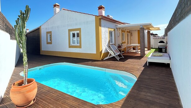 Un área junto a la piscina con un deck de madera, una pileta pequeña, una planta de cactus y un edificio con fachada amarilla y blanca.