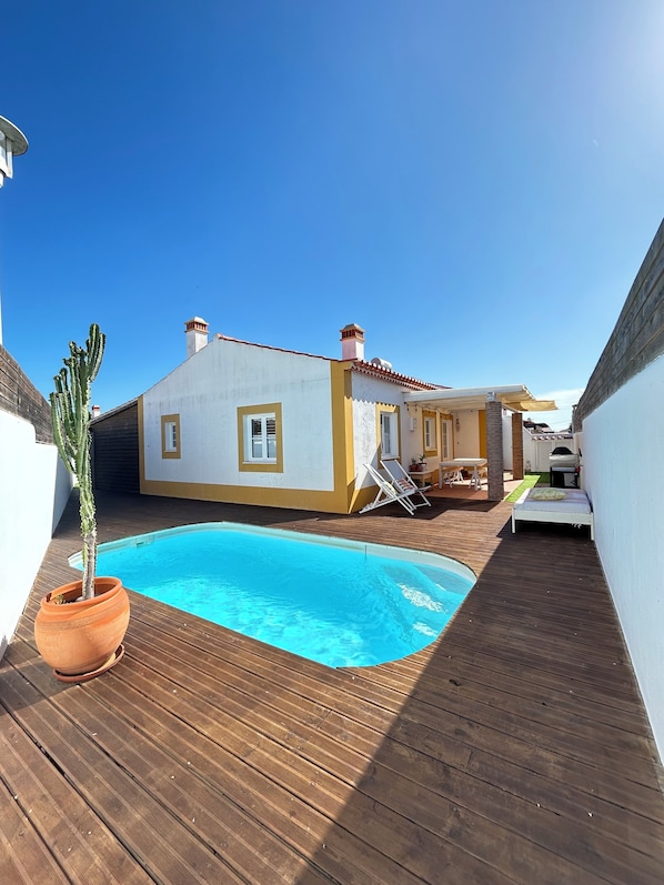Un espace aménagé au bord de la piscine, avec une terrasse en bois, une petite piscine, un cactus et un bùtiment à la façade jaune et blanche.