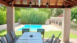 Une terrasse extérieure couverte avec une piscine, entourée de verdure et délimitée par une clôture.
