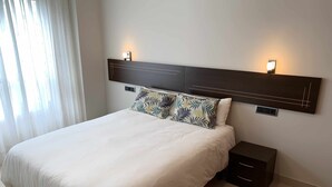 A neatly made bed with a white duvet, a dark headboard with two wall-mounted lights, and a small bedside table with a drawer.