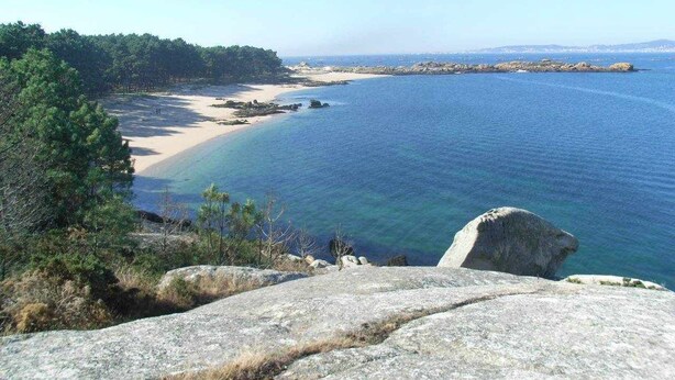 A coastal landscape with clear blue waters, sandy beach, and rocky outcrops.