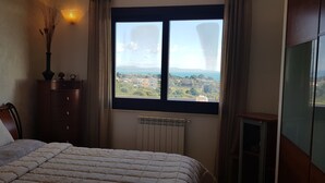 Un dormitorio con cama, cómoda, radiador y ventana con vistas a un pueblo y montañas.