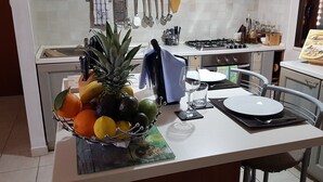 Una cocina con una mesa de comedor para dos, un frutero con naranjas y una piña, y un libro de cocina sobre la encimera.