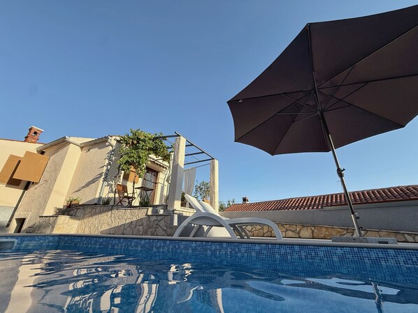 A swimming pool with a clear blue sky, a patio with a table and chairs, and a large umbrella.
