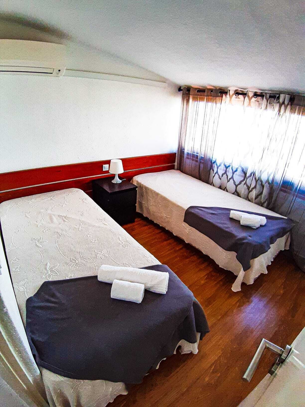Quarto com duas camas, mesa de cabeceira com abajur e janela com cortinas.