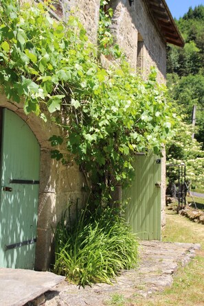 Un edificio de piedra con puerta y portón verdes, rodeado de vegetación y un camino de piedra.