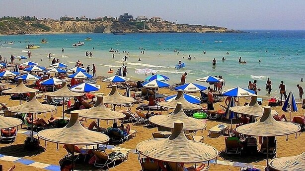 Ein Strand mit vielen Sonnenschirmen und Liegestühlen, ein klares blaues Meer und eine felsige Küste im Hintergrund.