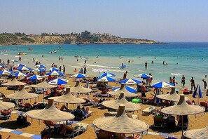 A beach with numerous sun umbrellas and beach chairs, a clear blue sea, and a rocky coastline in the background. - Excellent Holidays Rental Apartment, Free WiFi, High quality Complex. (Paphos)