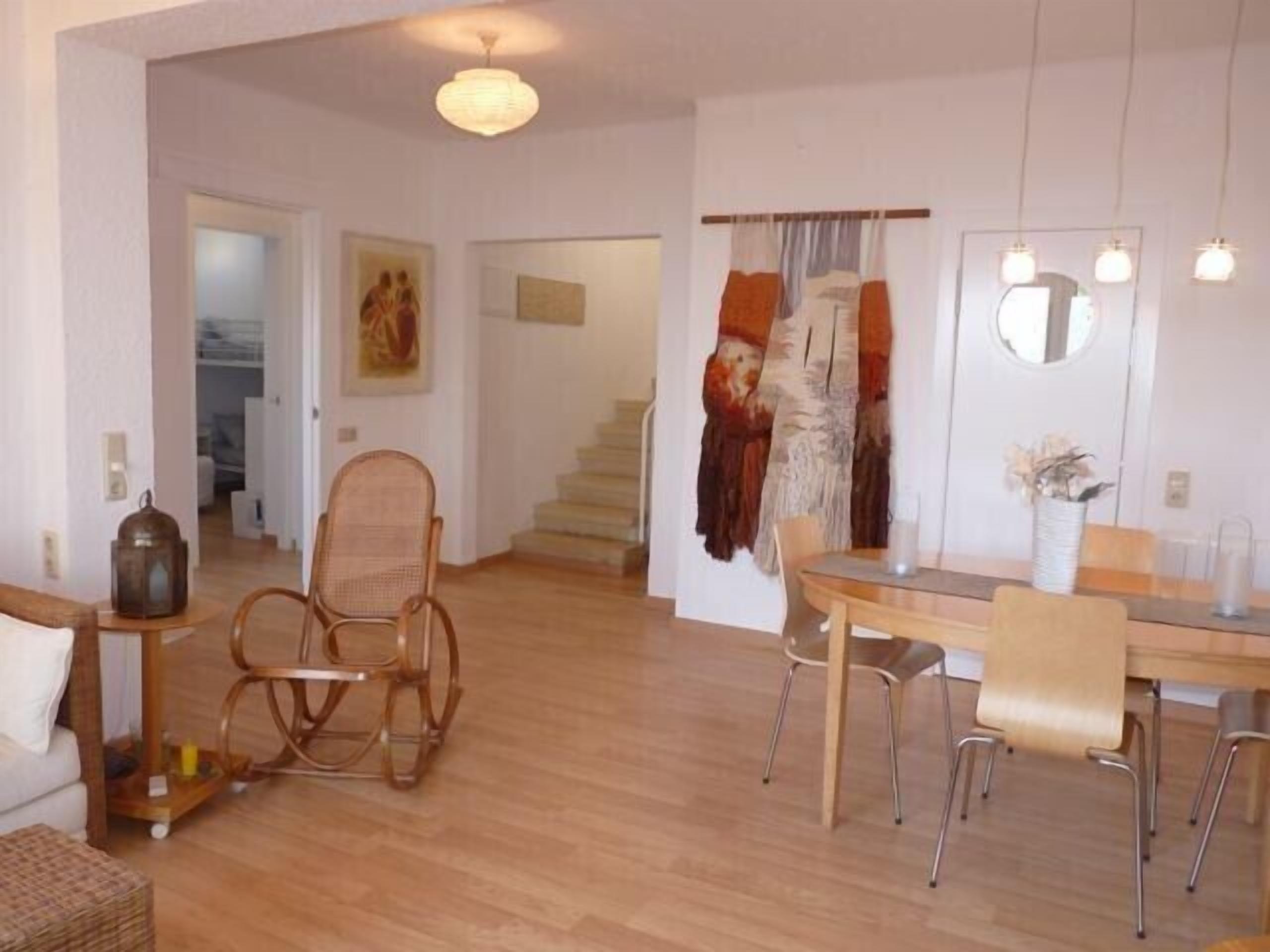 A spacious living area with a wooden floor, a dining table with chairs, a wicker chair, a sofa, and a lamp.