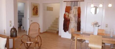 A spacious living area with a wooden floor, a dining table with chairs, a wicker chair, a sofa, and a lamp.