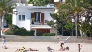 Une plage où des gens se prélassent au soleil et se baignent. On aperçoit un bâtiment doté d’un balcon, et des palmiers en arrière-plan.