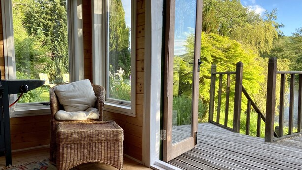 Une chaise en osier avec un coussin, placée près d’une grande fenêtre, un tapis à motifs et une terrasse en bois dotée d’une rambarde.