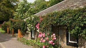 Un bâtiment rustique avec une façade en pierre, une clôture en bois et un jardin où poussent des rosiers roses.