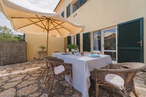 Ein Essbereich im Freien mit einem Tisch für zwei Personen, Korbstühle, einem großen Sonnenschirm und Blick auf ein Gebäude mit Fensterläden.