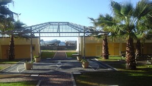 A covered walkway leading to a building with a glass roof, surrounded by palm trees and potted plants. - TWO-ROOM APARTMENT NEW SEA VIEW GALLINARA ISLAND DIRECTLY ON THE BEACH (ALBENGA)