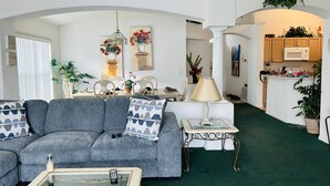Ein Wohnzimmer mit grauem Sofa, einem Glastisch und einer weißen Lampe. Im Hintergrund sind Pflanzen, ein Gemälde und eine Küche zu sehen.