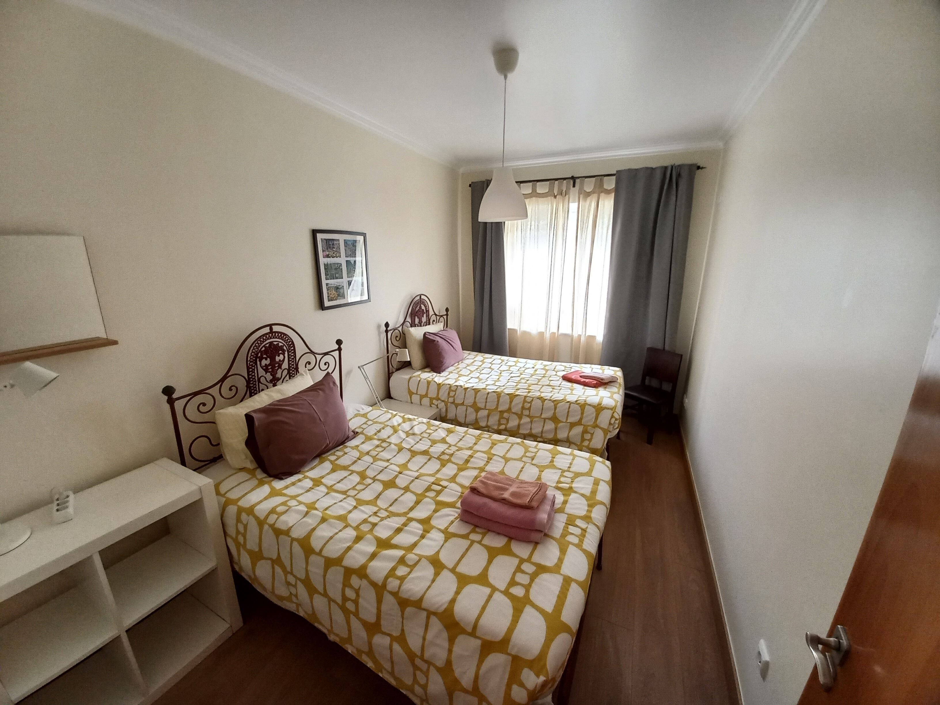 A small, well-lit room with two beds, a desk, and a chair.
