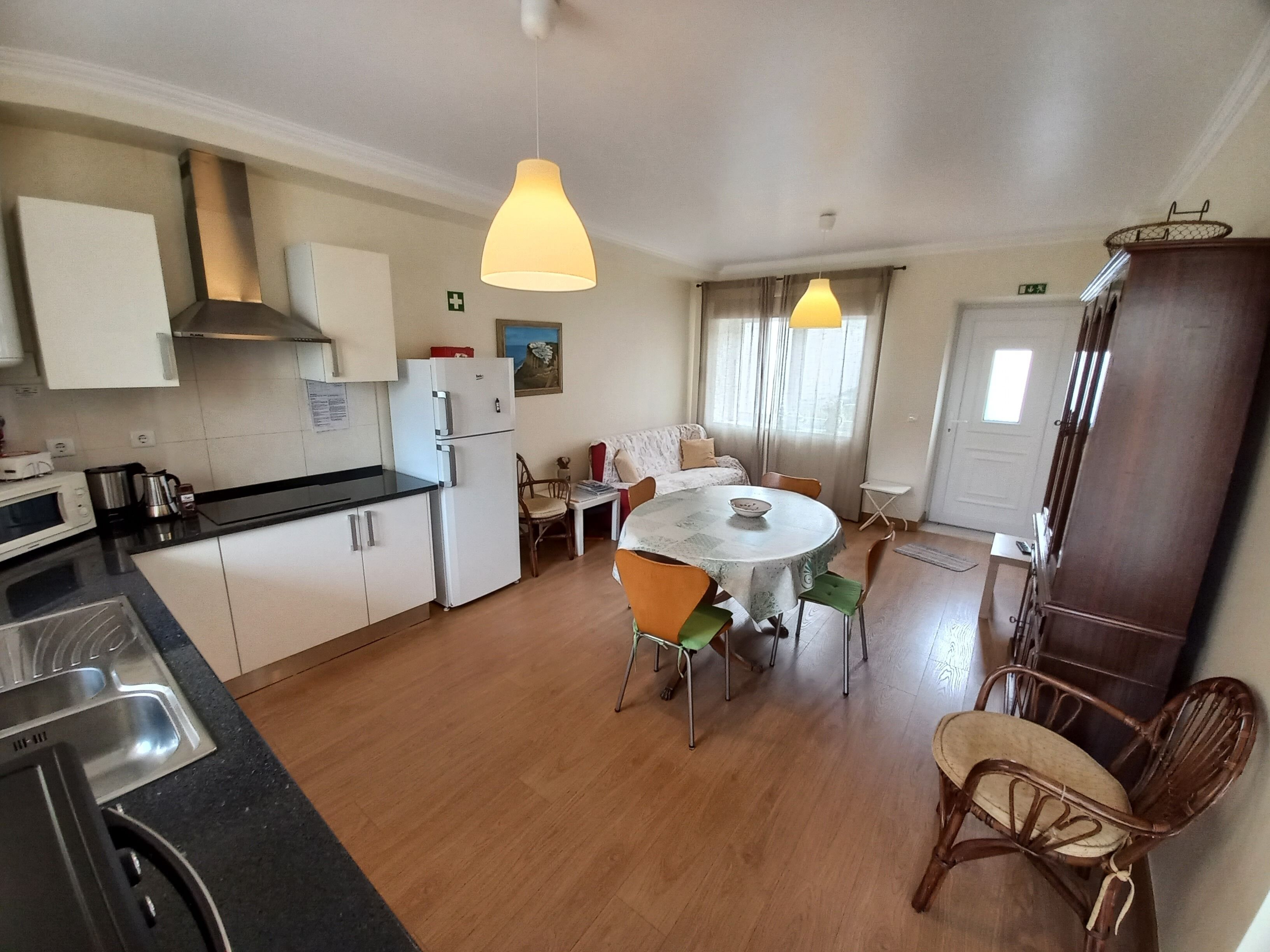 A compact kitchen with white cabinets, a refrigerator, a microwave, and a sink. There's a dining area with a round table and chairs, and a wooden cabinet to the right.