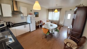 Eine kleine Küche mit weißen Schränken, einem Kühlschrank, einer Mikrowelle und einem Spülbecken. Es gibt einen Essbereich mit einem runden Tisch und Stühlen, und einen Holzschrank zur Rechten.
