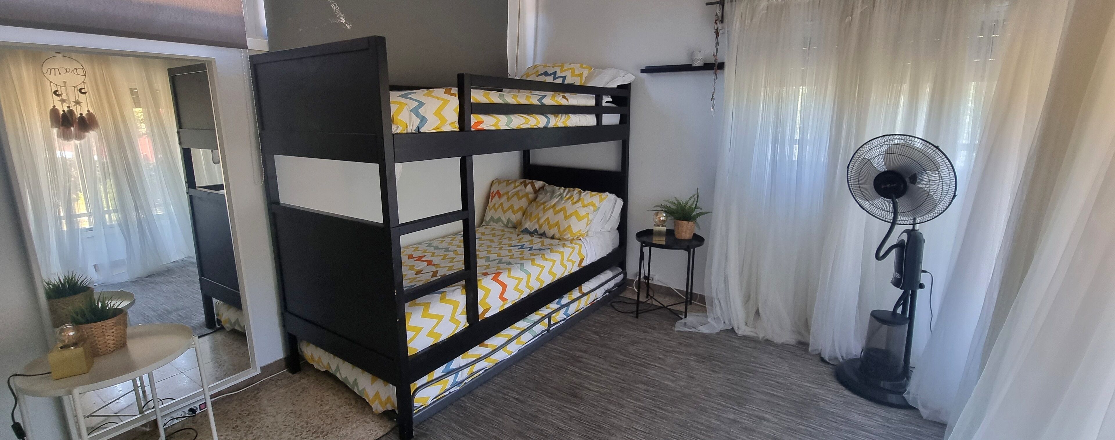 Una cama litera con ropa de cama estampada, una mesita con una planta, un ventilador de pie y una ventana con cortinas translúcidas.