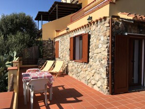 Un bâtiment en pierre doté d’une terrasse carrelée, de deux volets en bois et d’une table recouverte d’une nappe à carreaux.