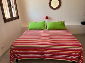 A bed with a striped bedspread and two green pillows, a red lamp, and a wall-mounted mirror.