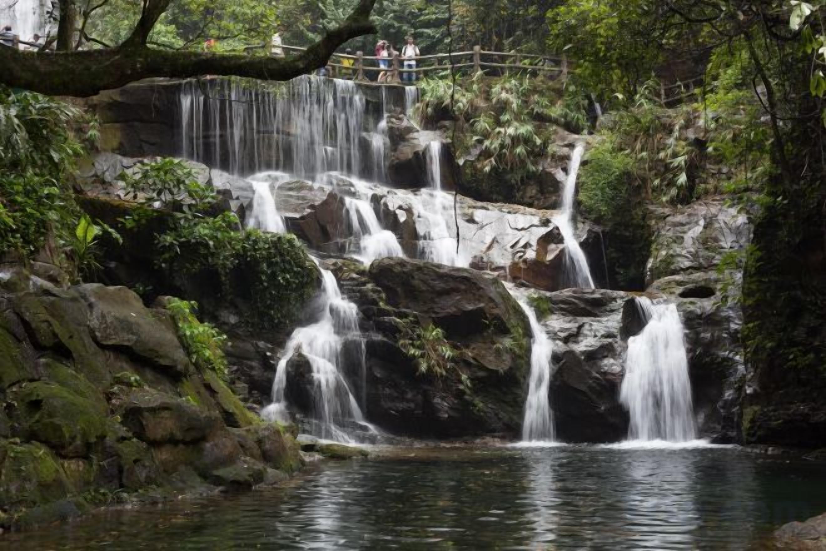 Ein mehrstufiger Wasserfall mit einer Holzbrücke, umgeben von üppigem Grün.