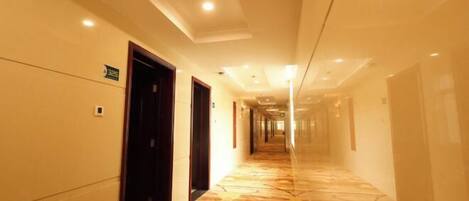 A long, narrow hotel corridor with a patterned floor, doors on the left, and a bright ceiling with recessed lighting.