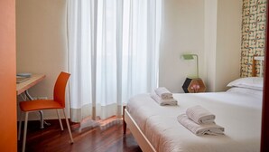 A hotel room with a bed, a desk with a chair, a window with sheer curtains, and a bedside table with a lamp and a vase. - TrastevereHome - Garibaldi Rome (Roma)