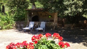 Un espace extérieur aménagé avec des sièges en pierre, comprenant deux fauteuils et une petite structure en pierre, qui pourrait être un barbecue ou un four, le tout entouré de verdure.