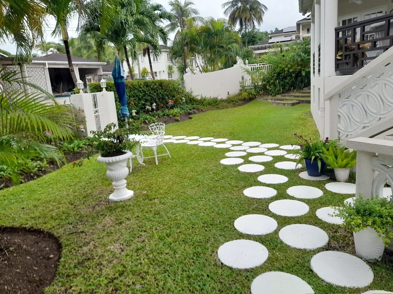 Un jardí ben cuidat amb un dibuix circular de pedres per a camí, una zona d'estar exterior blanca i vegetació abundant.