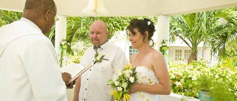 A couple in wedding attire under a white gazebo with a view of lush greenery and a building in the background.