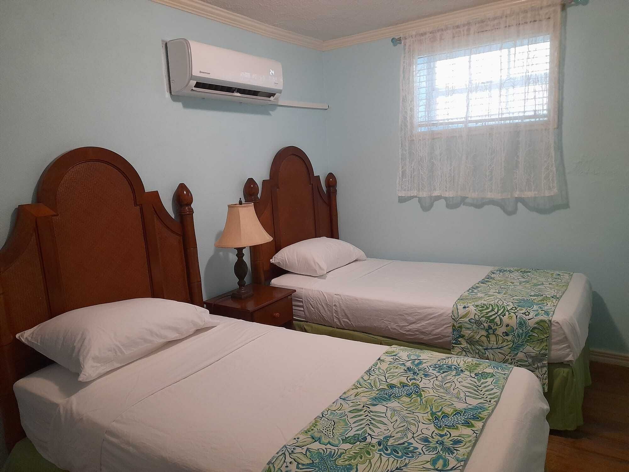 A room with two beds, a nightstand with a lamp, an air conditioner, and a window with a lace curtain.