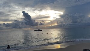 Ein Strand mit ruhigem Wasser, ein Segelboot und eine Person im Wasser.