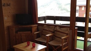 Een gezellige kamer met houten details, een televisie op een lage plank, een bank, een kleine tafel met twee rode objecten en een uitzicht op een groen landschap door grote ramen.