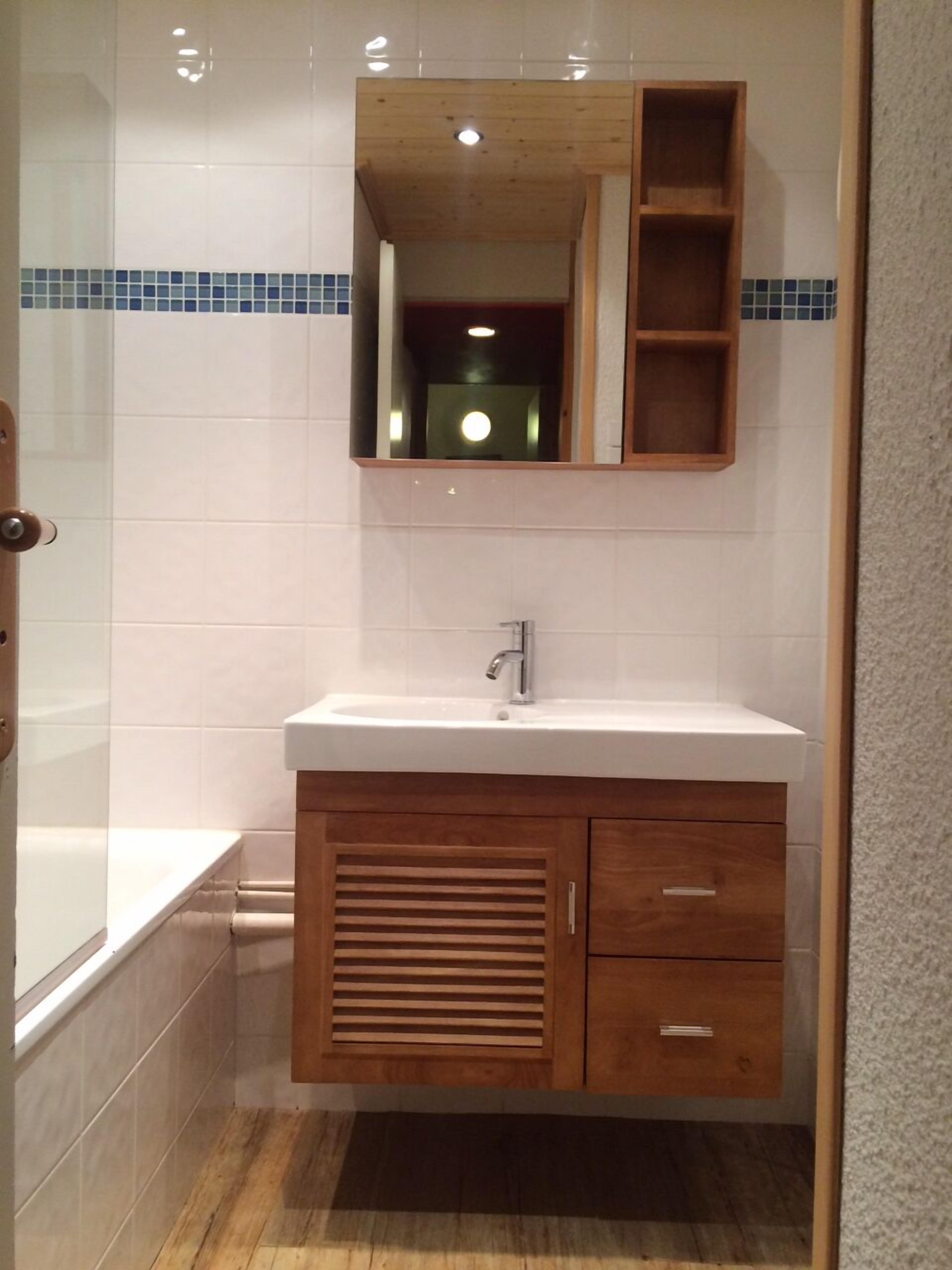 A bathroom with a wooden vanity, a white sink, and a mirror above it. There is a shower area to the left and a door leading to another room.