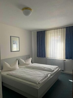 Une chambre d’hôtel avec deux lits, un luminaire au plafond, une fenêtre avec des rideaux et un tableau encadré au mur.