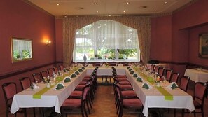 Une salle à manger élégante, avec de longues tables dressées pour le repas, de grandes fenêtres ornées de rideaux et un lustre.