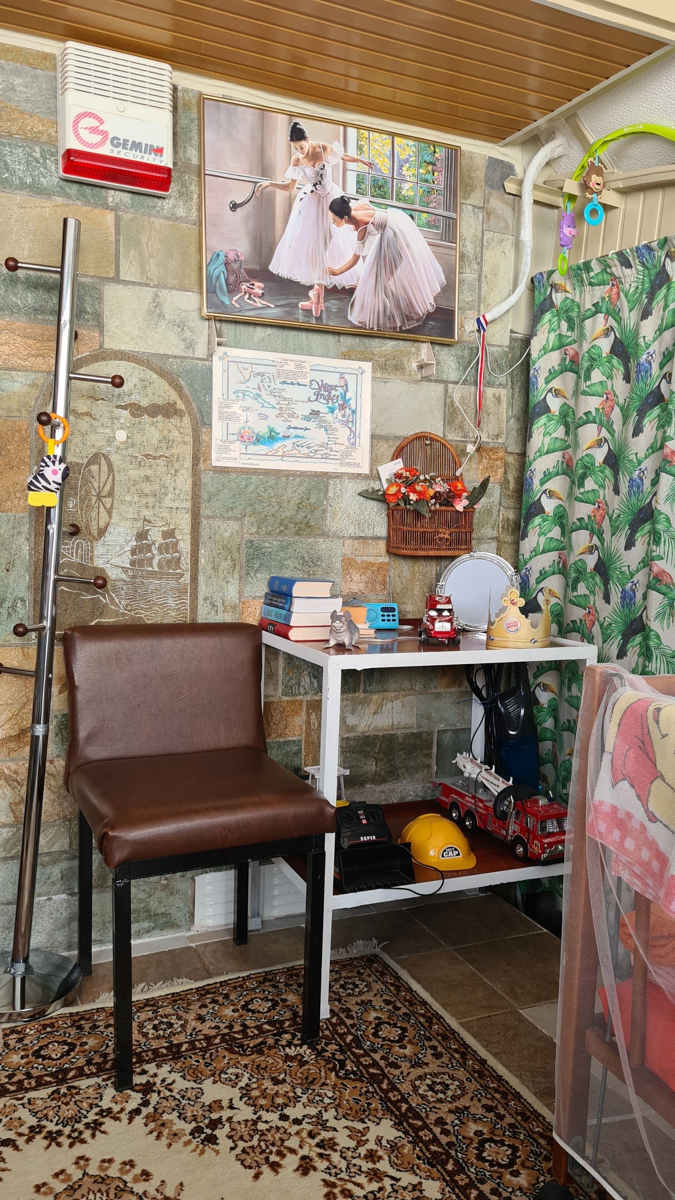 A room with a brown leather chair, a metal desk with various items, a framed picture of ballerinas, a security device, and a tropical curtain.