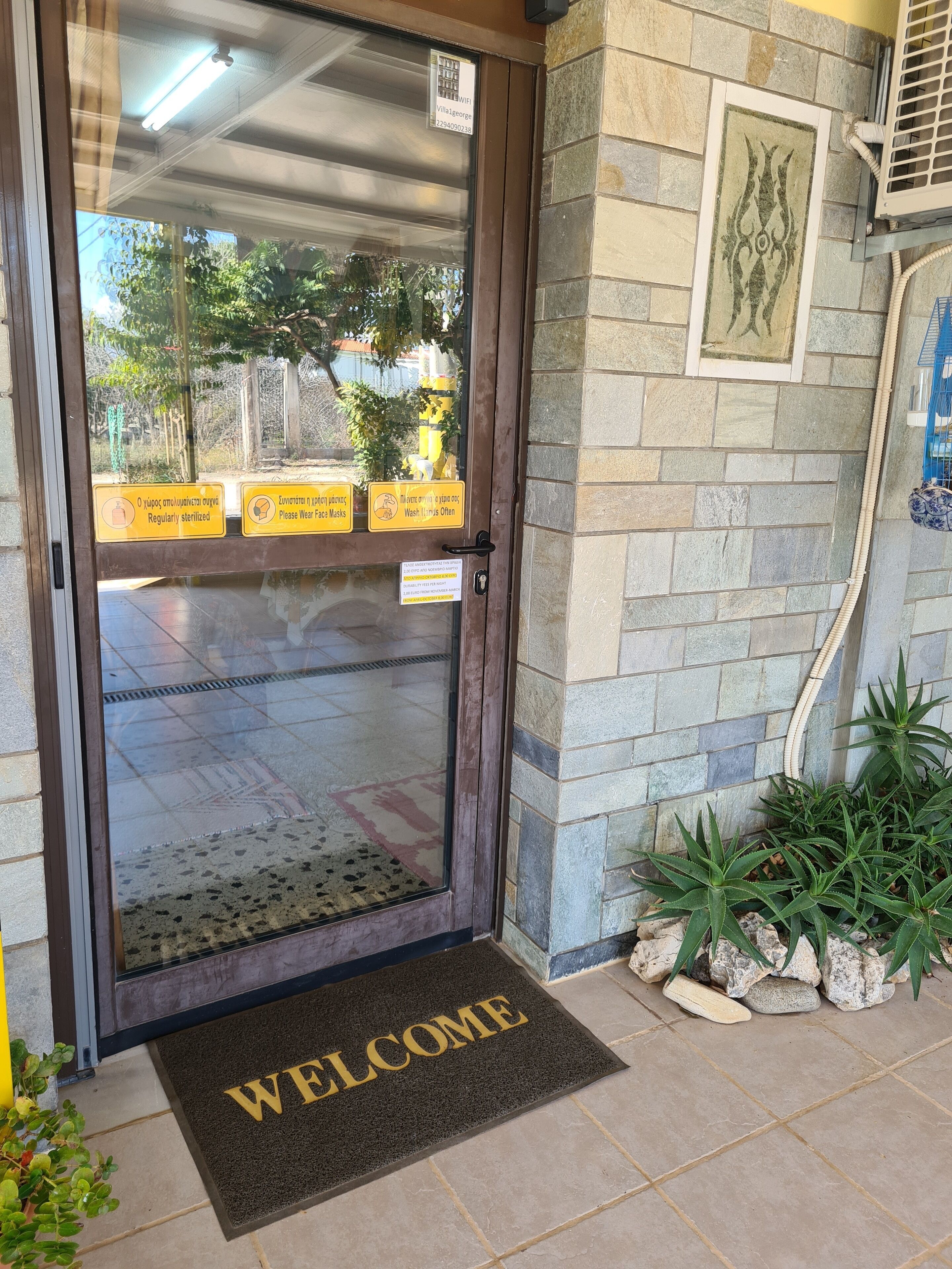 A hotel entrance with a glass door, a welcome mat, and signs advising to wear a face mask and wash hands often.