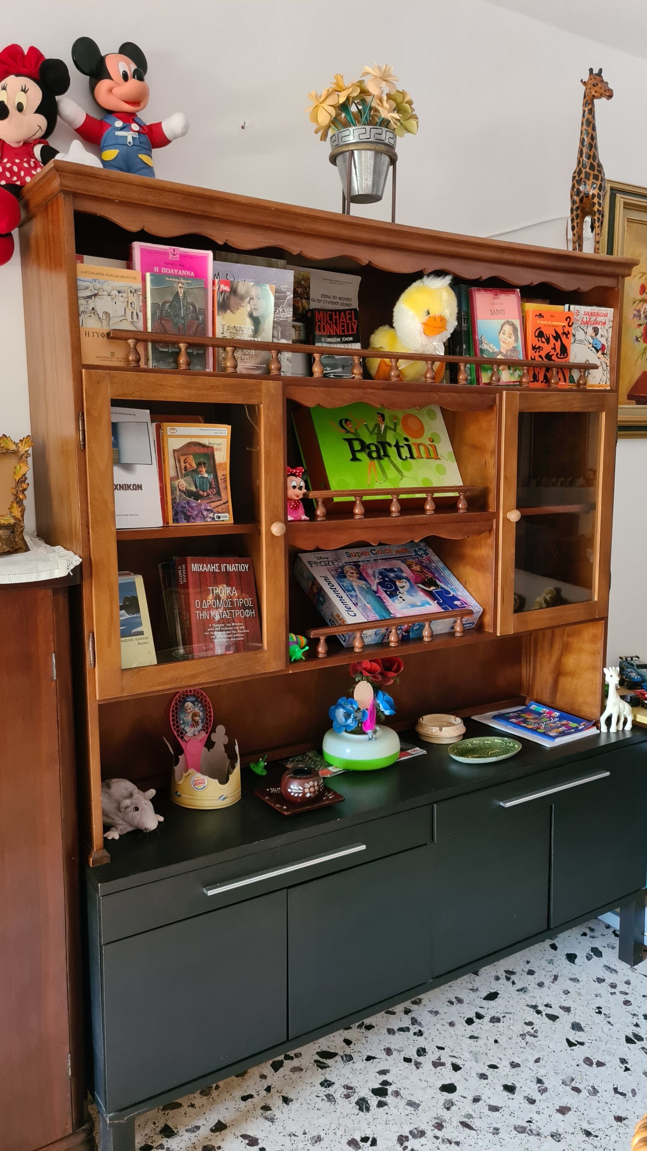 A wooden bookshelf with various books, a Mickey Mouse plush, a duck plush, and a small figurine.