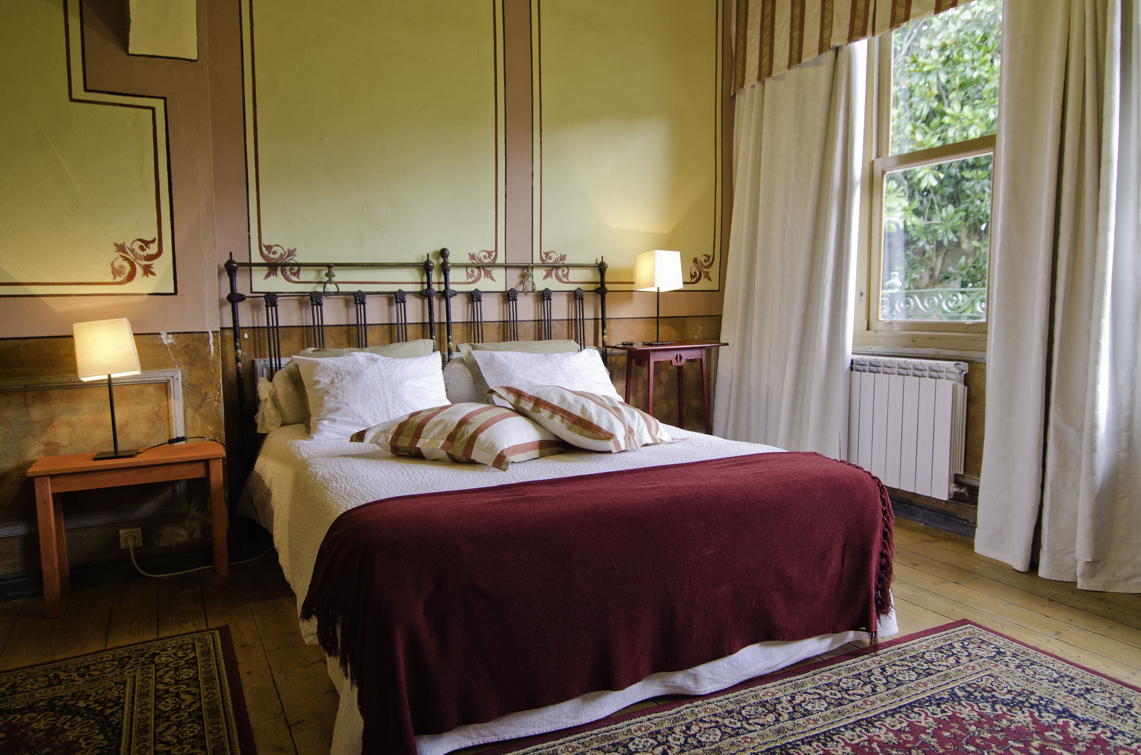 Een keurig opgemaakt bed met een rode sprei en twee kussens, een nachtkastje met een lamp, en een raam met gordijnen.