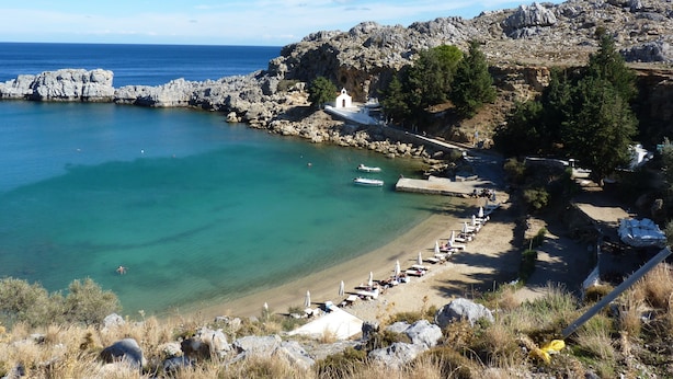 Una spiaggia costiera con terreno roccioso, acque turchesi limpide e una fila di ombrelloni.