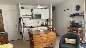 Et kompakt køkken med hvide skabe, en mikrobølgeovn og en vask. Der er et spisebord med stole og et lille rundt bord med en stol.