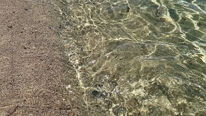Una platja amb aigües poc profundes i cristal·línes, pedres i una muntanya a la distància, juntament amb algunes persones banyant-se.