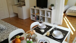 Una zona de menjador amb una taula preparada per a dues persones, incloent plats, coberts i un bol de fruita. Al fons, es veuen un microones i una televisió.