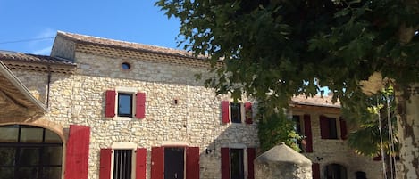 A stone building with red shutters, a stone well, and a grassy courtyard.