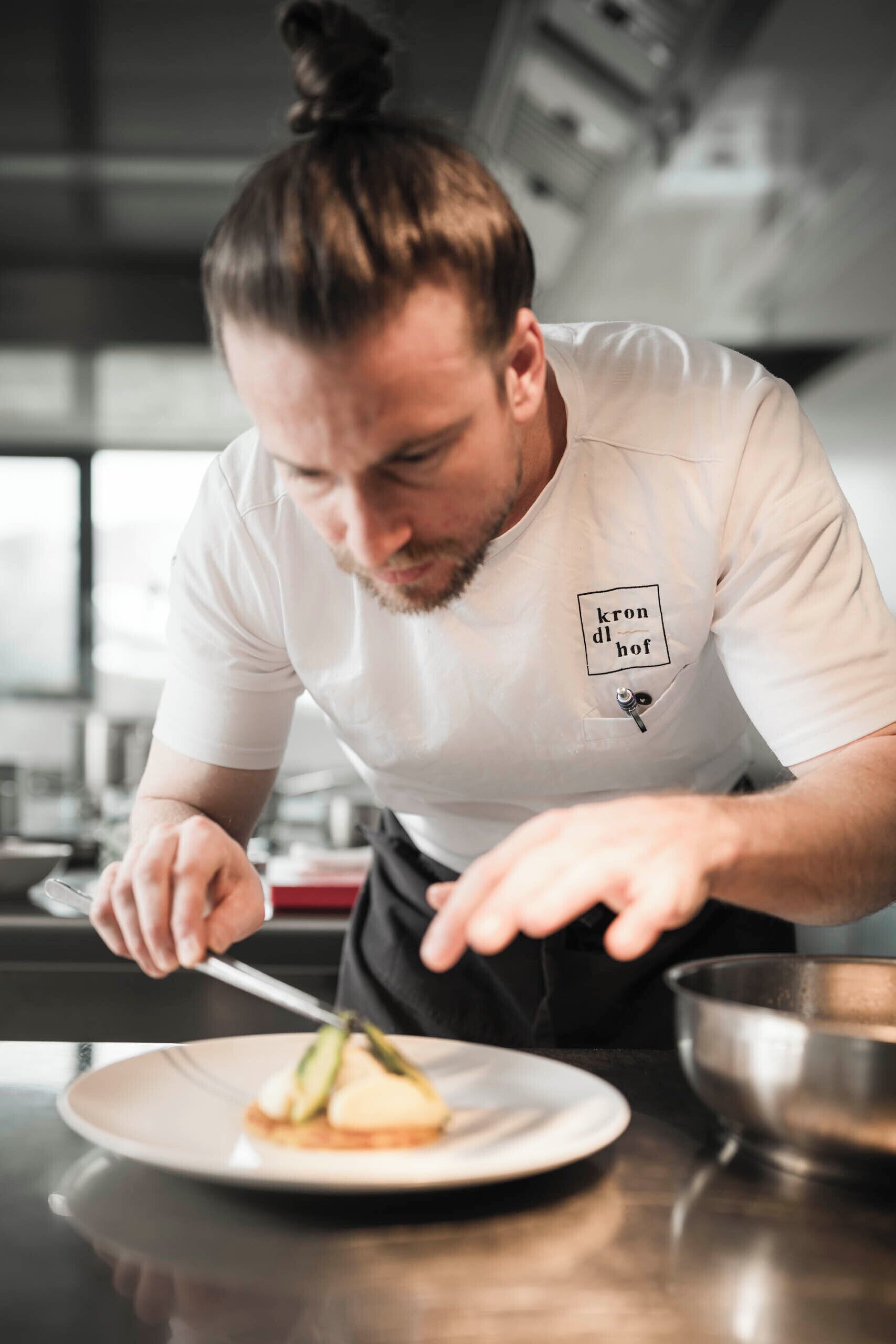 A chef in a white shirt with a logo, preparing food in a kitchen with stainless steel appliances.