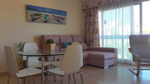 A living room with a glass table, white chairs, a sofa, a potted plant, and a painting of boats on the wall.